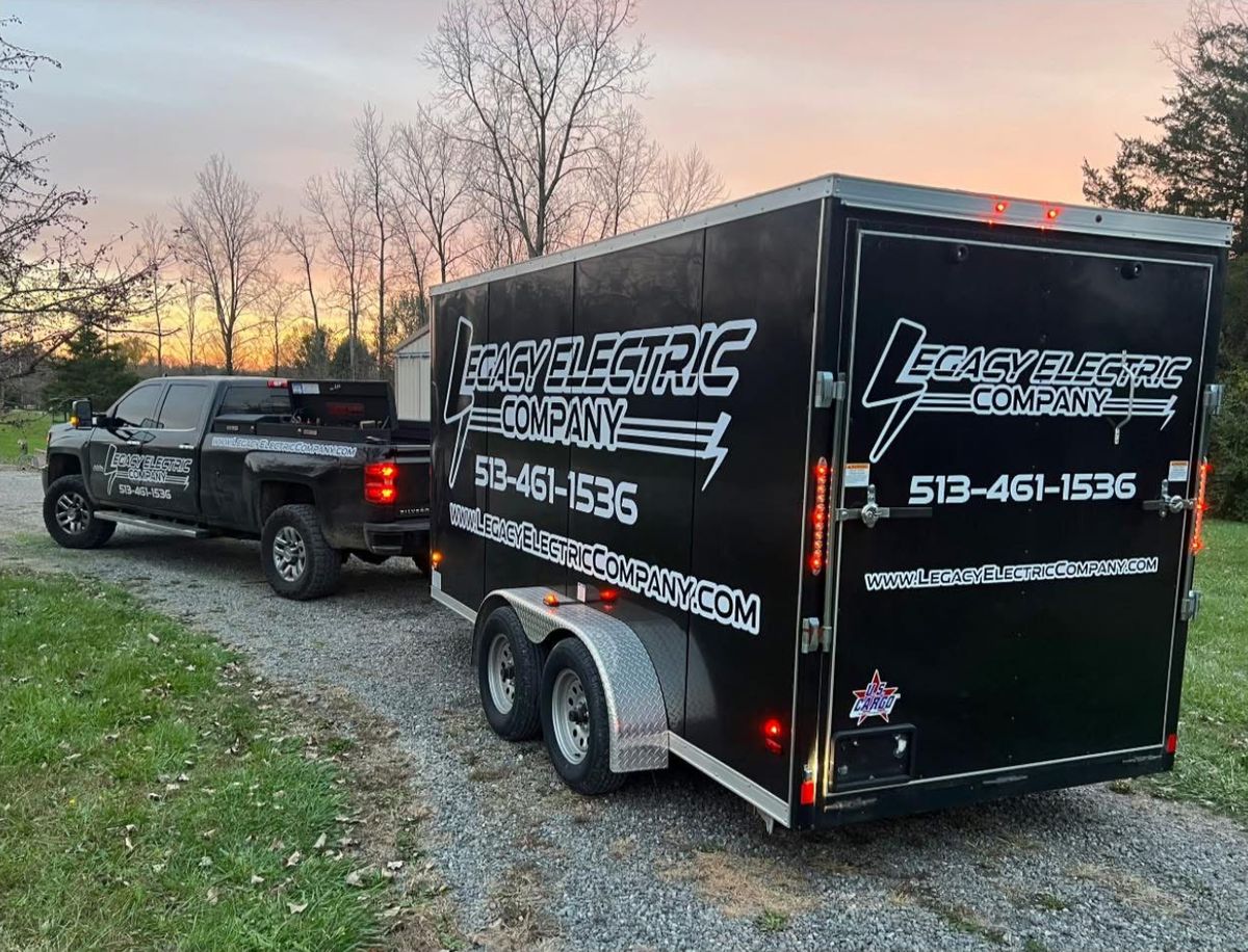 Legacy Electric Company's black pickup truck towing the company's black enclosed work trailer, lettered with the Legacy Electric Company logo and 513-461-1536, on a gravel drive at sunset